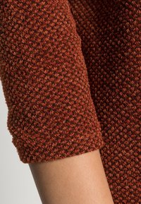 Texturovaná pletenina v rezavé barvě s krátkým rukávem zakončeným manžetou. Materiál je na dotek měkký, s mírně nerovným povrchem a jemnými odstíny barev.