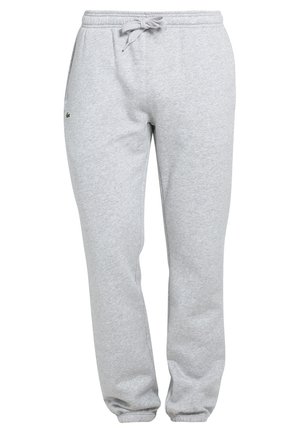 Mann trägt ein dunkelblaues T-Shirt, hellgraue Jogginghose mit kleinem Logo auf der Tasche und weiße Sportschuhe, steht mit einer Hand in der Tasche.
