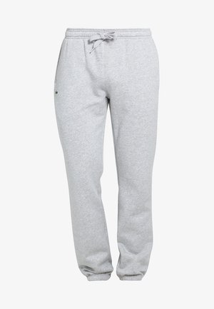 Man draagt een marineblauw t-shirt, lichtgrijze joggingbroek met een klein logo op de zak en witte sportschoenen, staat met één hand in de zak.