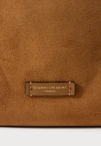 Pungă din suede maro cu o etichetă din piele cu litere aurii embossate pentru "GIANNI CHIARINI FIRENZE," asigurată prin cusături de un bej deschis.