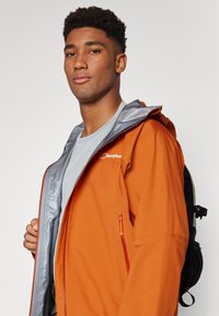 Veste imperméable orange avec une capuche grise, fermeture éclair sur le devant et poche latérale. Portée par-dessus une chemise grise, accompagnée d'un sac à dos noir.
