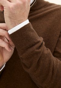 Brauner Strickpullover mit einer strukturierten Oberfläche, ausgestattet mit einem gerippten Bündchen und einem weißen Innenkragen. Die Passform ist eng und betont die Kontur des Arms.