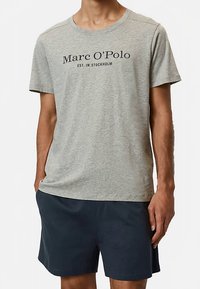 Camiseta gris de manga corta con texto negro que dice "Marc O'Polo EST. IN STOCKHOLM", combinada con pantalones cortos azul marino. Tejido suave, diseño casual.