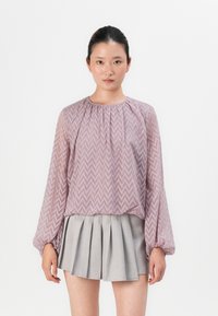 Light purple, zigzag-patterned blouse with long, tapered sleeves and gathered neckline, worn with a gray pleated skirt. Backdrop is plain white.