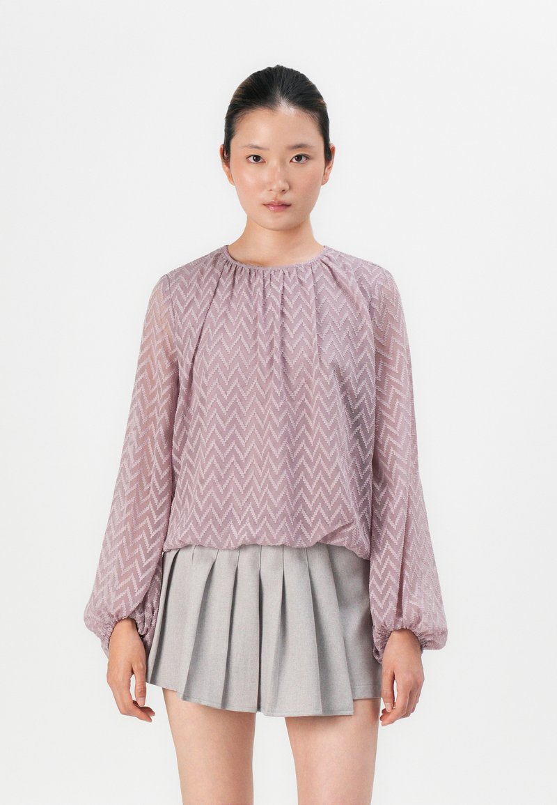 Light purple, zigzag-patterned blouse with long, tapered sleeves and gathered neckline, worn with a gray pleated skirt. Backdrop is plain white.