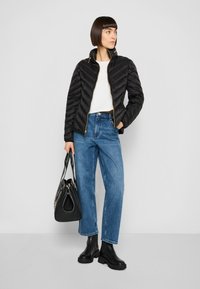 Zwarte pufferjas met een chevron-doorstikt ontwerp, gecombineerd met een lichtgekleurde top, blauwe wijde jeans en een zwarte schoudertas. Zwarte laarzen.