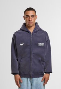 Sudadera con capucha y cremallera en algodón azul oscuro, que presenta bolsillos frontales y gráficos impresos en blanco en el pecho. Diseño informal con un corte relajado.