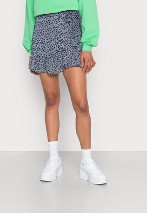Vrouw met een marineblauwe bloemen minirok, witte platform sneakers, witte sokken en een felgroene longsleeve top, staand tegen een witte muur.