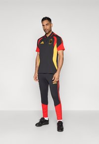 Conjunto atlético em preto, vermelho e amarelo, apresentando uma camisola polo com colarinho e calças com seções inferiores vermelhas, logótipo e riscas. Sapatos pretos.