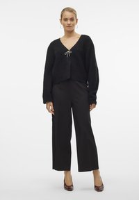 Pull en maille texturée noir avec un col en V et un détail de nœud à l'avant, associé à un pantalon noir ajusté et des chaussures à talons marron.