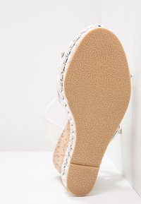 Sandale compensée avec semelle en caoutchouc beige texturée, garniture tressée argent et blanche, et talon compensé à motif liège sur fond blanc.