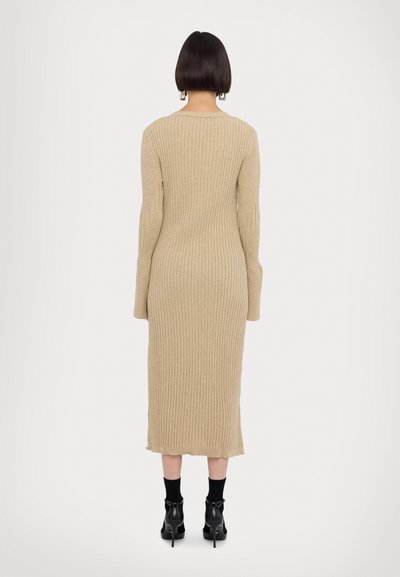 Beige gebreide jurk met ribbelstructuur, lange mouwen en een rechte pasvorm die net onder de knie eindigt. Gecombineerd met zwarte enkellaarsjes en sokken.