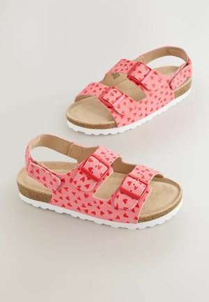 Rosa sandaler med hjärtatmönster, två justerbara remmar, korkfotbädd och vit gummisula. Öppen baksida för enkel på- och avtagning.