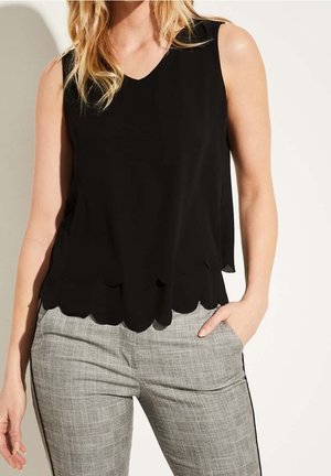 Kobieta w czarnej, bezrękawnikowej bluzce z falowanym dołem i szarych spodniach w kratę, stojąca z jedną ręką w kieszeni na jasnym tle.