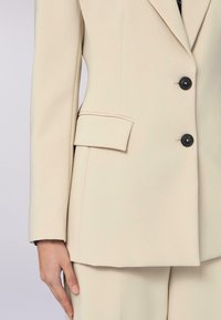 Beige op maat gemaakte blazer met een gladde textuur, twee zwarte knopen en een klepzak. Het ontwerp heeft een gestructureerde silhouette en scherpe revers.