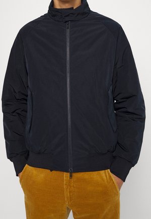 Mężczyzna ubrany w czarną kurtkę zapinaną na zamek z kieszeniami oraz musztardowożółte spodnie sztruksowe, z rękami w kieszeniach, na jasnym tle.