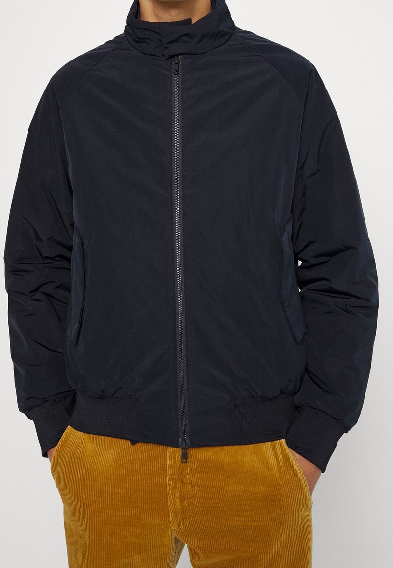 Homme portant une veste zippée noire avec des poches et un pantalon en velours côtelé jaune moutarde, les mains dans les poches, devant un fond clair.