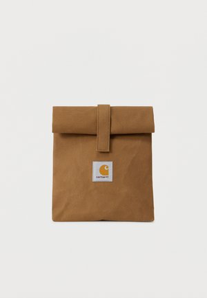 Bolsa de almuerzo de lona marrón con la parte superior doblada asegurada por una correa y un parche de logo cuadrado en el centro delantero.