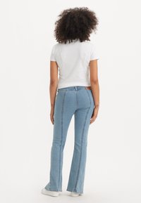 Vaqueros acampanados de color azul claro con detalles de costura lateral. El modelo lleva una camiseta blanca ajustada y se ven unas zapatillas blancas en la parte inferior.