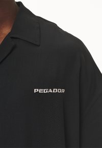 Maglietta nera a maniche corte con colletto. Presenta il logo ricamato "PEGADOR" in argento sul lato sinistro. Tessuto liscio e leggero.