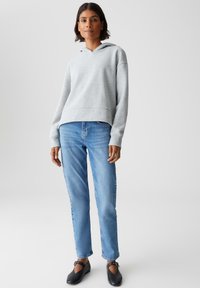 Giovane donna che indossa un hoodie grigio chiaro, jeans azzurri chiari e scarpe Mary Jane nere, in piedi contro uno sfondo chiaro e uniforme.