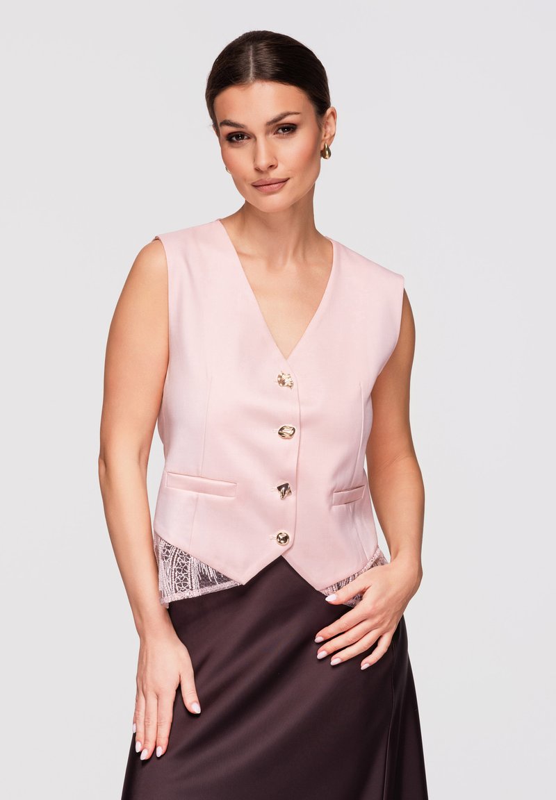 Femme aux cheveux foncés portant un gilet sans manches rose pâle à boutons dorés sur une jupe en satin foncé, posant devant un fond clair.