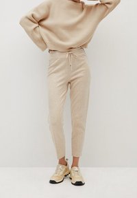 Béžový žebrovaný svetr s kulatým výstřihem, doplněný krémovými zúženými kalhotami s elastickým pasem a stahovací šňůrkou, a světlými teniskami.