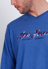 Camisa de manga larga azul con escote en V hecha de una tela texturizada que presenta un logo en rojo, blanco y azul con el texto "Lois Jeans" y "Original Garments".