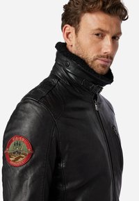 Zwarte leren jas met een zachte, gestructureerde kraag, ritssluiting en een rode geborduurde patch op de linkerschouder.