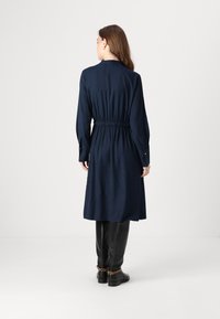 Mørkeblå kjole med elastisk midje, lange ermer, og krage. Kjolen går under kneet, og den er parret med høye svarte støvler.