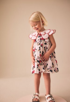 Jong meisje met kort blond haar dat een witte jurk draagt met Minnie Mouse prints en een grote geschulpte kraag, glimlachend en naar beneden kijkend.