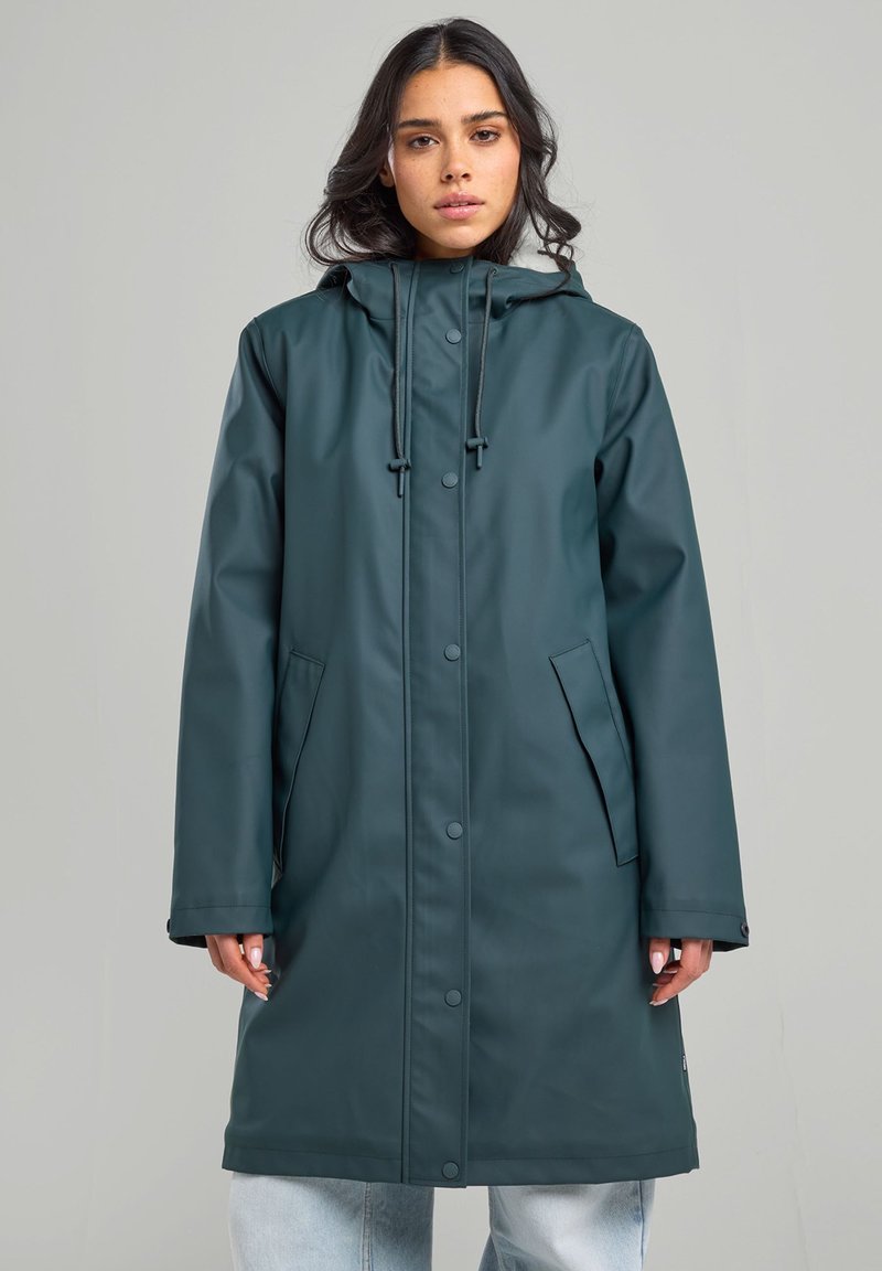Manteau imperméable vert foncé avec une capuche, des boutons-pression et deux poches latérales. Dispose d'un cordon de serrage ajustable au col et d'une longueur plus longue.