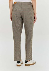 Osoba w brązowych, w kratkę spodniach i białych trampkach z czarnymi detalami, stojąca na tle jasnego, jednolitego tła.