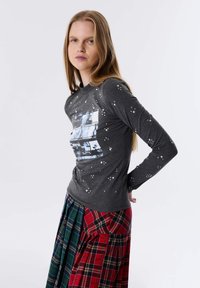 Grijs shirt met lange mouwen en fotografische print, versierd met zilverkleurige studs; gecombineerd met een geruite, meerkleurige rok met tinten rood en groen.