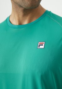 Kadrą priartinta vyro nuotrauka, vilkinčio žalią marškinėlį su apvaliu kaklu ir mažuoju Fila logotipu kairėje krūtinės pusėje.