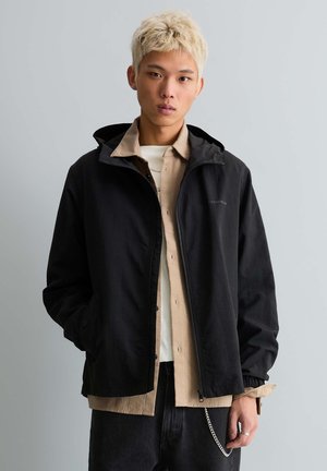 Ung mand med blondt hår iført en sort jakke over en beige skjorte med knapper og sorte bukser, stående foran en grå baggrund.