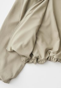 Tissu de veste beige montrant la doublure intérieure, la fermeture éclair en métal et le détail élastiqué de l'ourlet sur fond blanc.