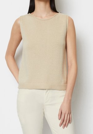 Vrouw die een mouwloos beige gebreid topje en lichtbeige broek draagt, staand tegen een effen witte achtergrond.