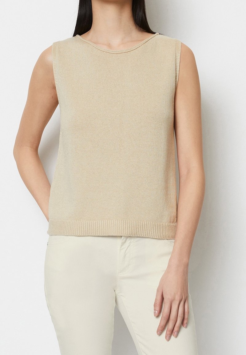 Femme portant un haut en tricot beige sans manches et un pantalon beige clair, debout devant un fond blanc uni.