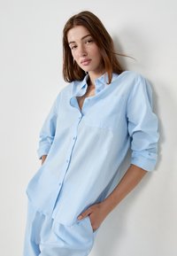 Joven mujer con cabello largo castaño que lleva una camisa de botones azul claro y pantalones a juego, de pie contra una pared blanca con una mano en el bolsillo.