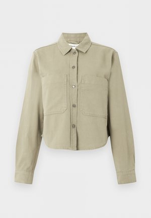 Beeži lühike jakk pikkade varrukatega, kraega, esikinnituse ja kahe suure rinnataskuga tavalise tausta ees.