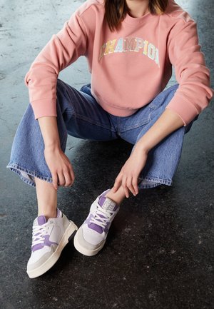 Persoon zit op de vloer met een roze "CHAMPION" trui, blauwe spijkerbroek en witte sneakers met paarse accenten.