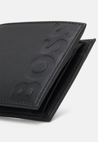 Juodas odinis piniginė su iškilniu "BOSS" logotipu, lygia tekstūra ir sulankstomu dizainu su matomais siūlėmis. Kompaktiška ir minimalistinė.