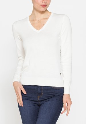 Donna che indossa un maglione bianco a scollo a V e maniche lunghe e jeans blu scuro, in piedi con una mano poggiata sulla coscia.