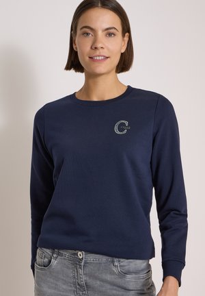 Ung kvinde med lige brunt hår iført en navyblå langærmet skjorte med et lille "C" logo på brystet og lysegrå jeans.