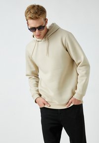 Sudadera beige con un bolsillo frontal, cordones y puños acanalados. Combinada con pantalones negros. Textura suave y lisa, diseño minimalista.