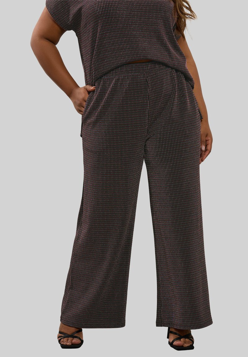 Vrouw in textuur brede pijpen broek en bijpassende top met één hand in haar zak, staande met de voeten in zwarte gekruiste sandalen.