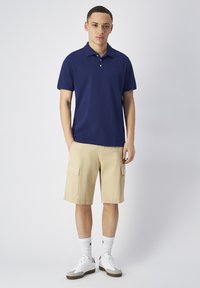 Granatowa koszulka polo z kołnierzykiem, beżowe szorty cargo z bocznymi kieszeniami, białe skarpetki i szaro-białe trampki. Gładkie tło.