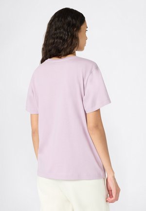 Donna con lunghi capelli mossi che indossa una semplice maglietta corta di colore viola chiaro e pantaloni avorio, mostrata di spalle su uno sfondo bianco.