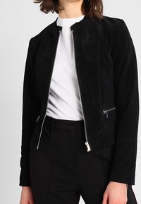 Černá semišová bunda na zip přes bílý tričko s kulatým výstřihem, k tomu černé kalhoty s vysokým pasem.